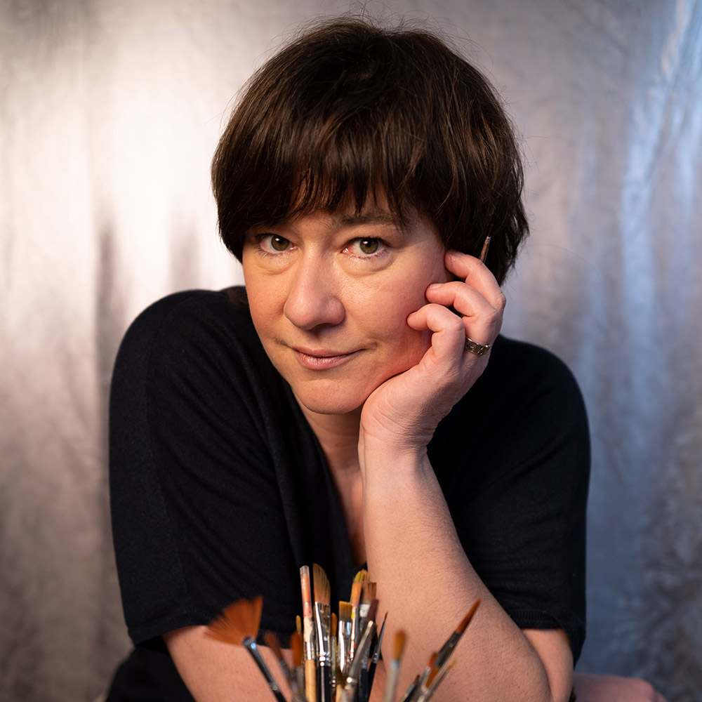 Portrait Nadine Jork, sie schaut auf eine Hand leicht gestützt mit einem Pinsel in dieser Hand direkt in die Kamera